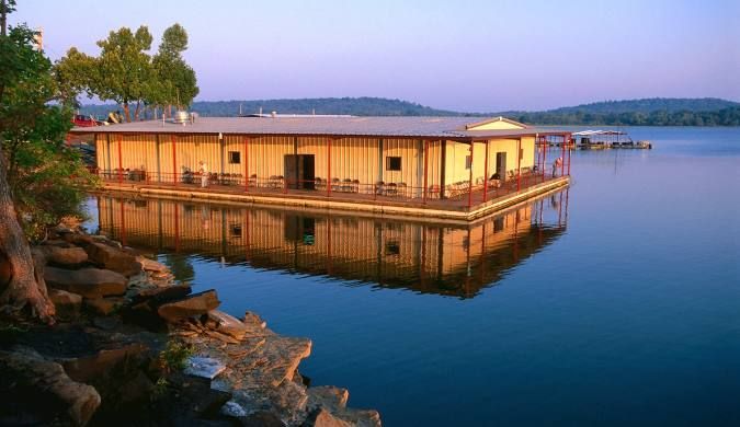 Floating wooden building on a lake, reflecting in the water during sunset. Trees and shore are in the foreground.