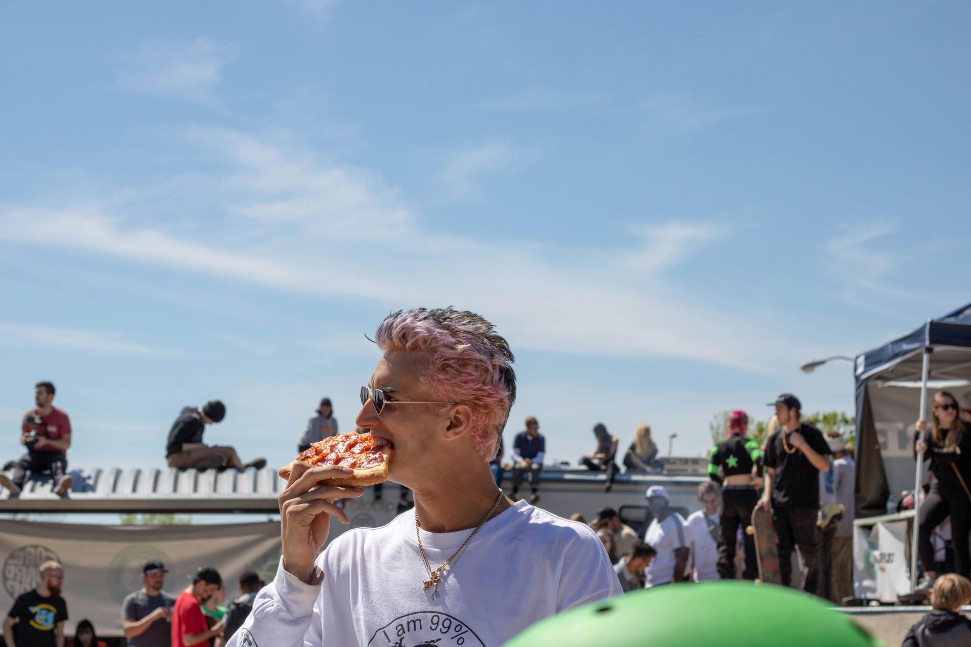 Man with pink-streaked hair eating pizza at skatepark, people watching in background, sunny day.