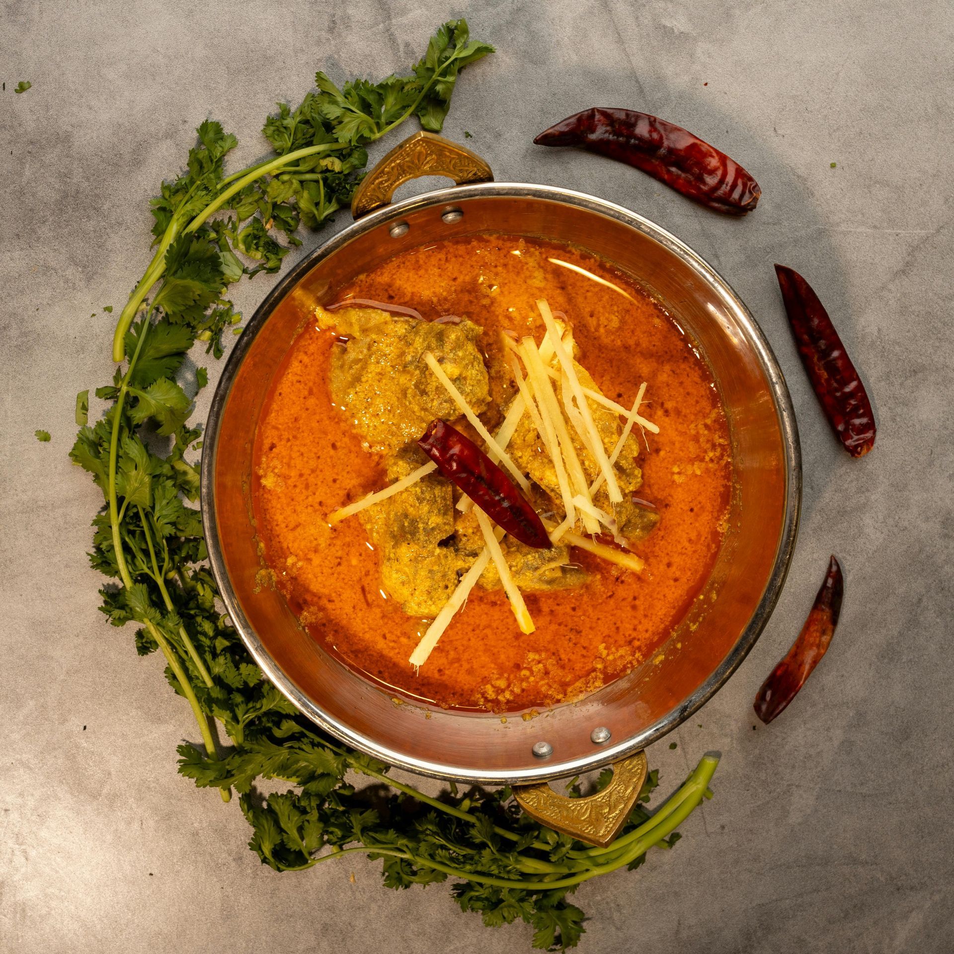 Bowl of orange curry with fish, garnished with ginger, chilies, and cilantro on a gray surface.