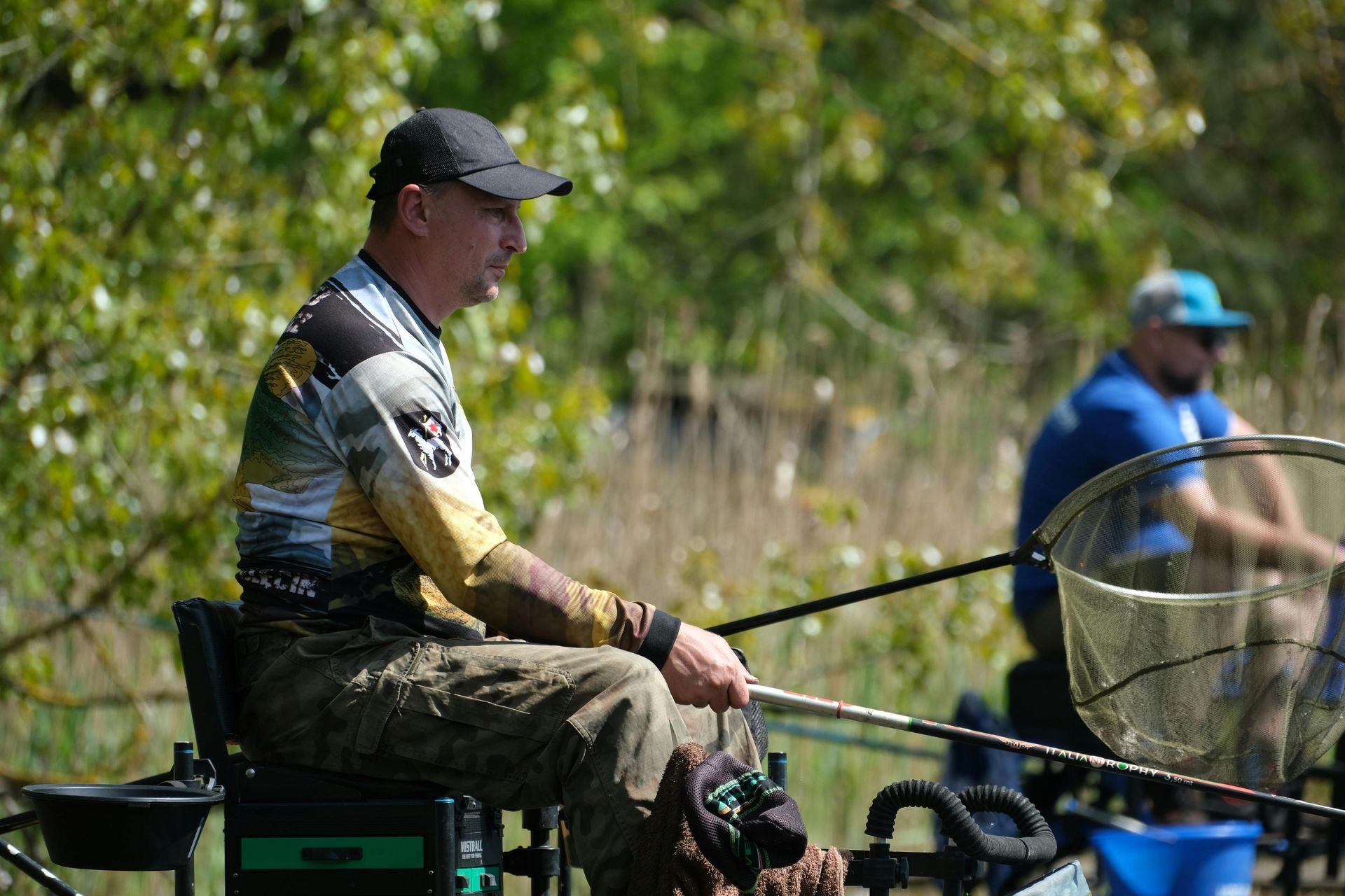 Man in fishing attire seated, holding a fishing rod. Another person fishing in the background, near water and foliage.