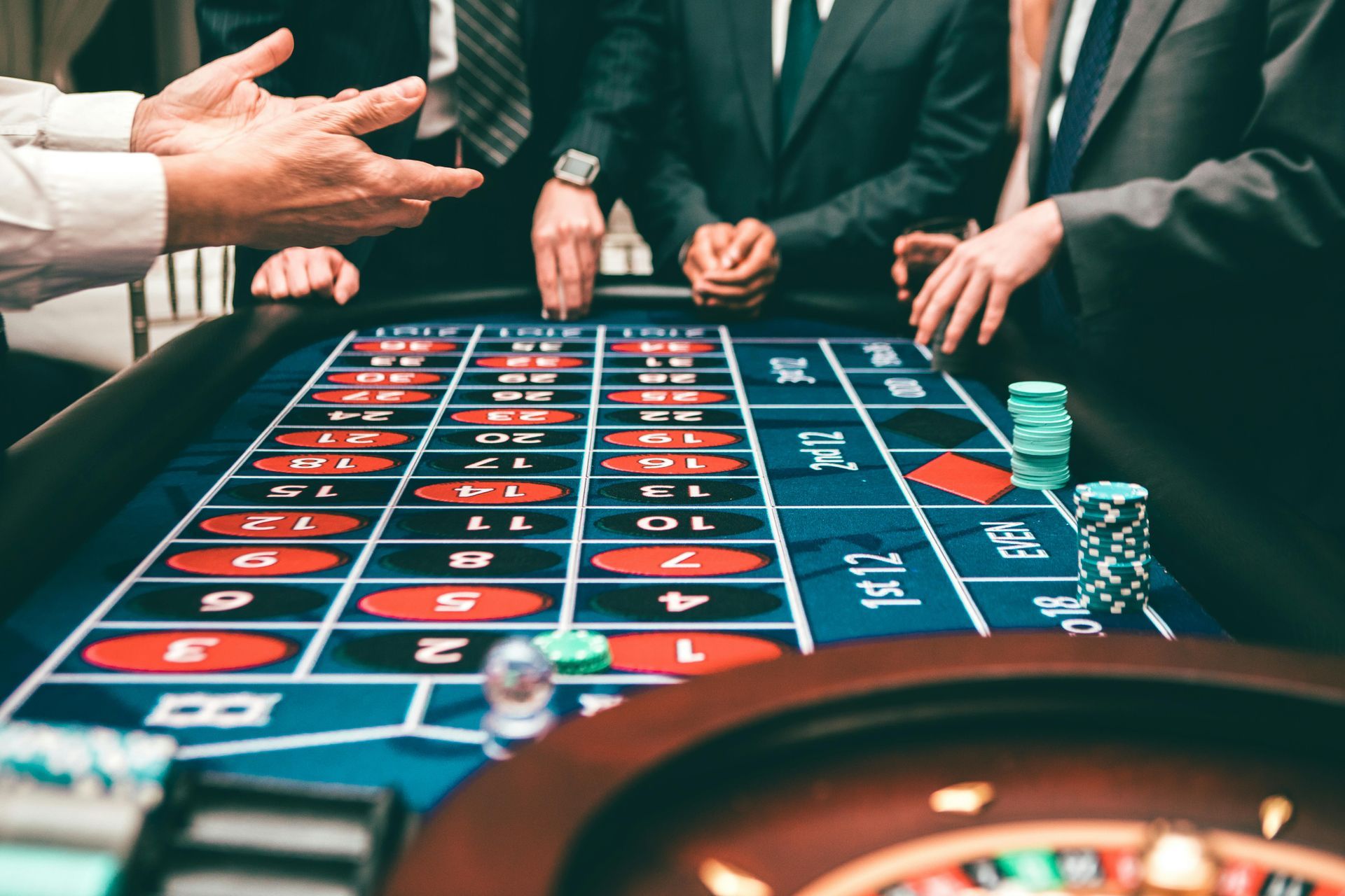 People at a roulette table in a casino, hands gesturing above the layout with chips and wheel visible.