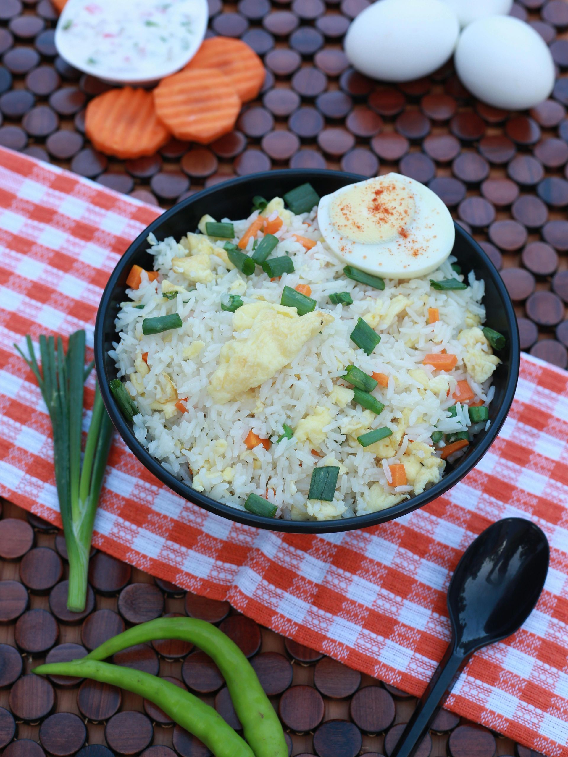 Egg fried rice in a black bowl, garnished with egg and vegetables, on a checkered cloth.