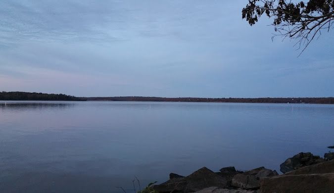 Calm lake reflecting a dusky blue sky; a few trees line the horizon.