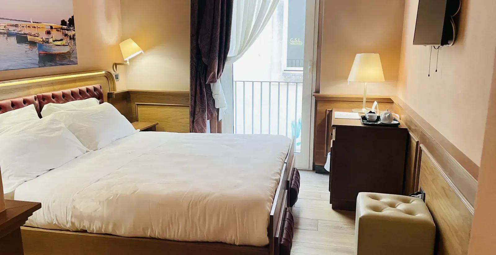 Camera d'albergo con letto, scrivania e balcone. Pareti rosa, finiture dorate e un dipinto con vista sull'acqua.