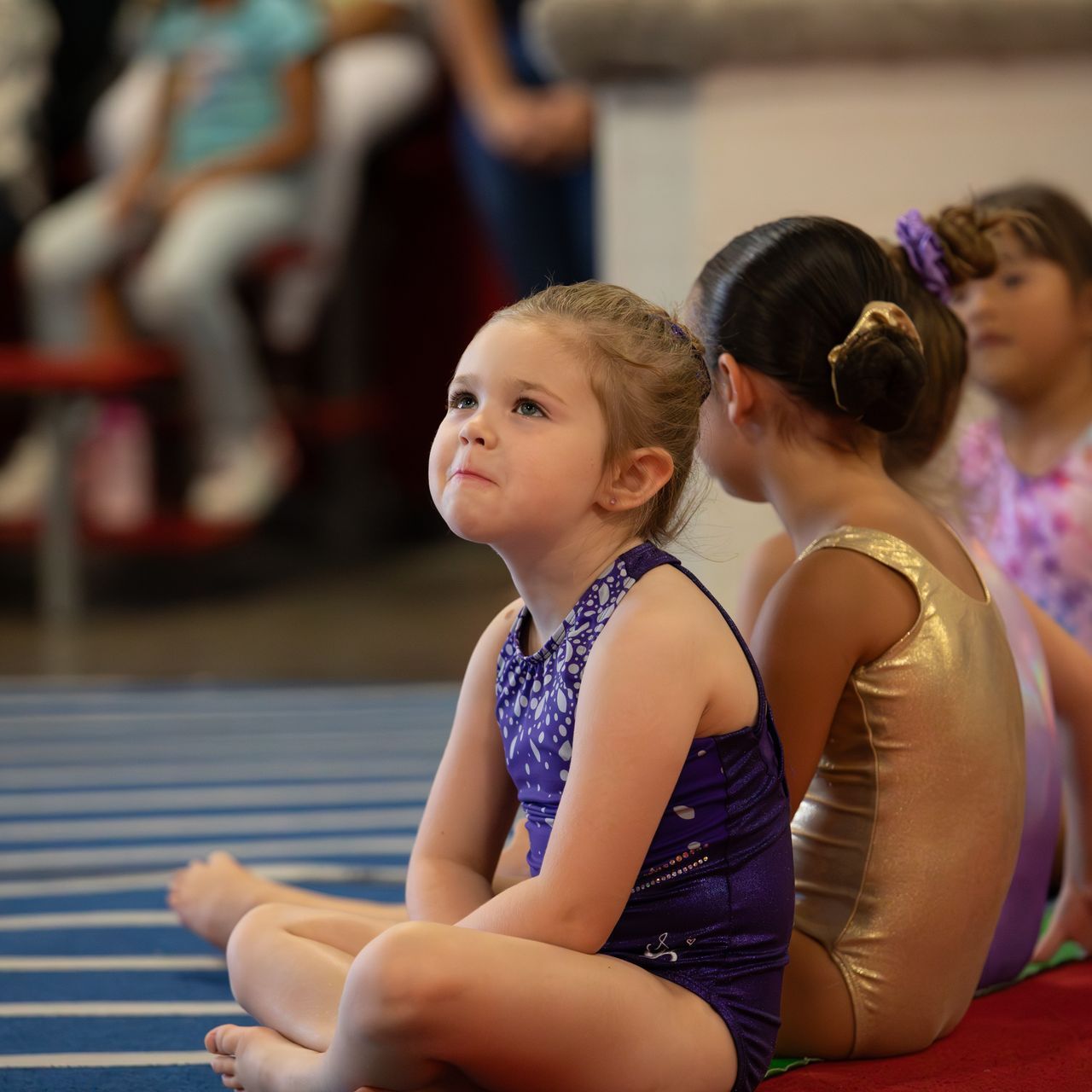 A group of young girls are sitting on the floor and one of them is wearing a purple leotard