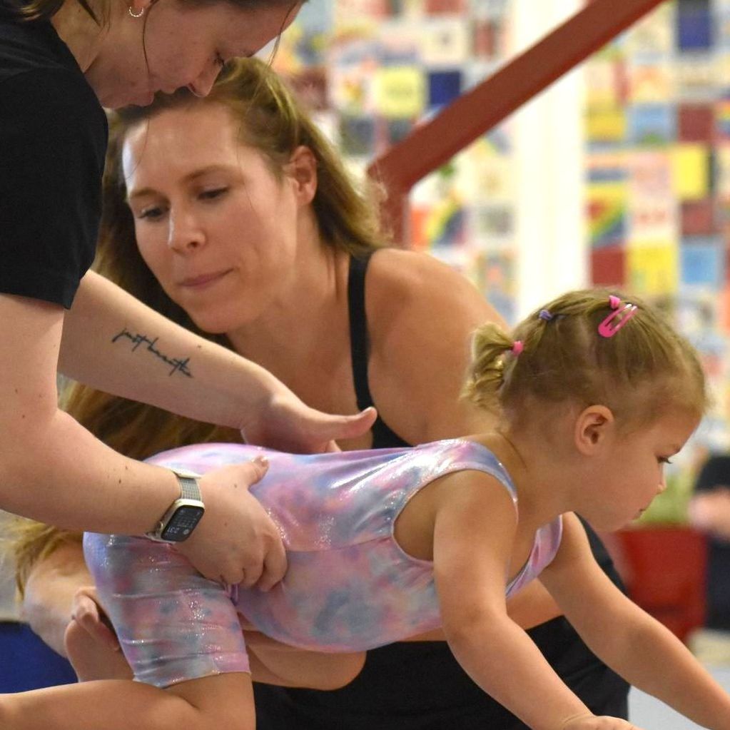 A woman with a tattoo on her arm is helping a little girl balance on a balance beam