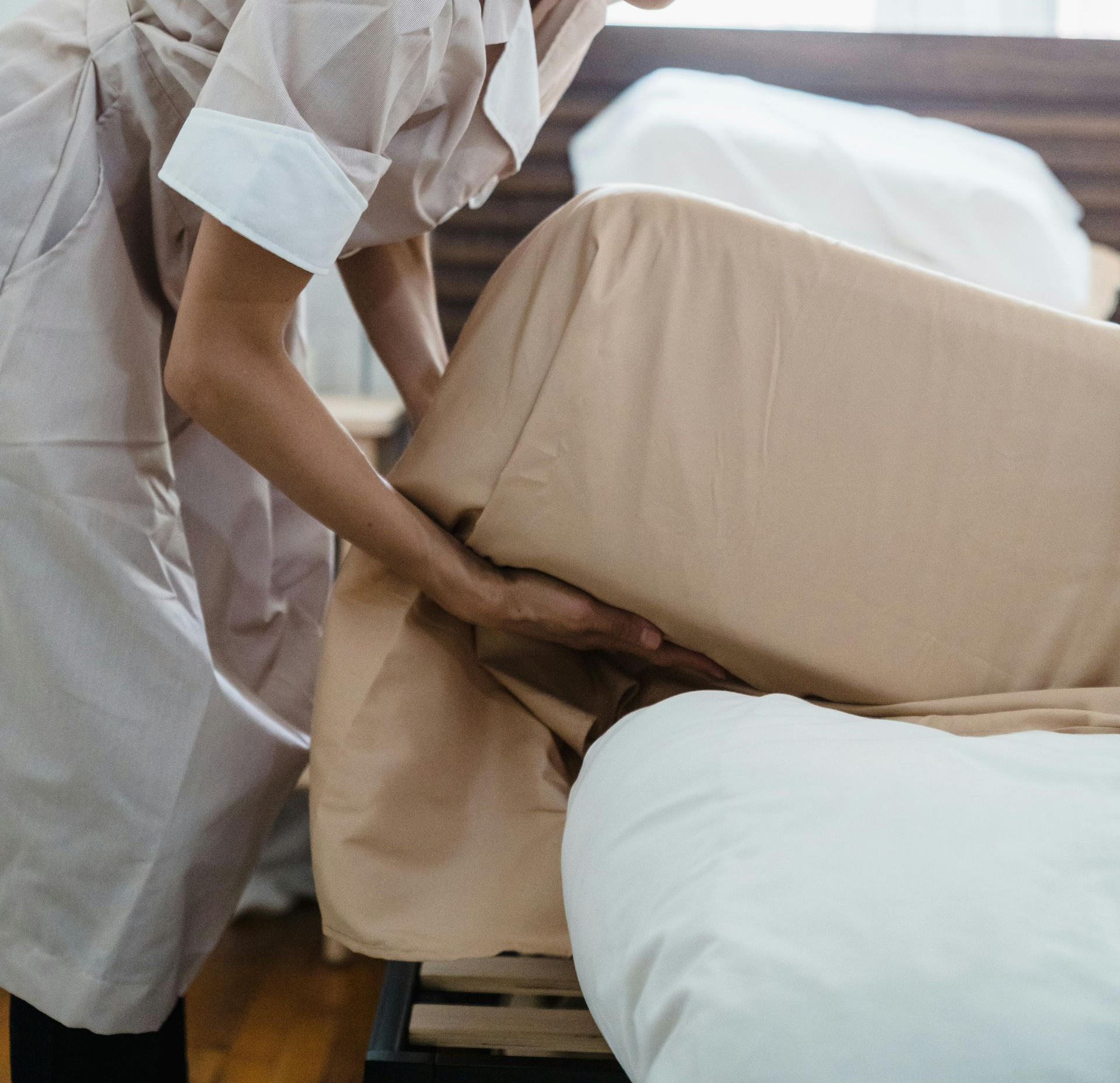 Person in uniform making a bed with tan sheets and white pillow.
