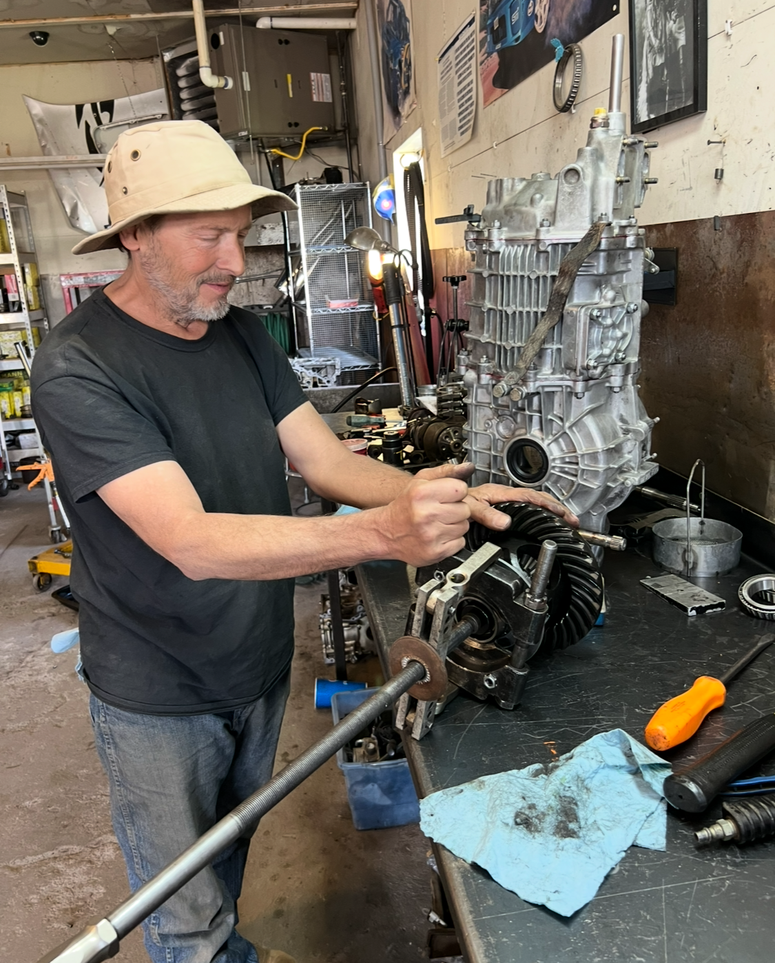 Mechanic points to an engine on a workbench. He wears a hat, black shirt, and is in a garage.