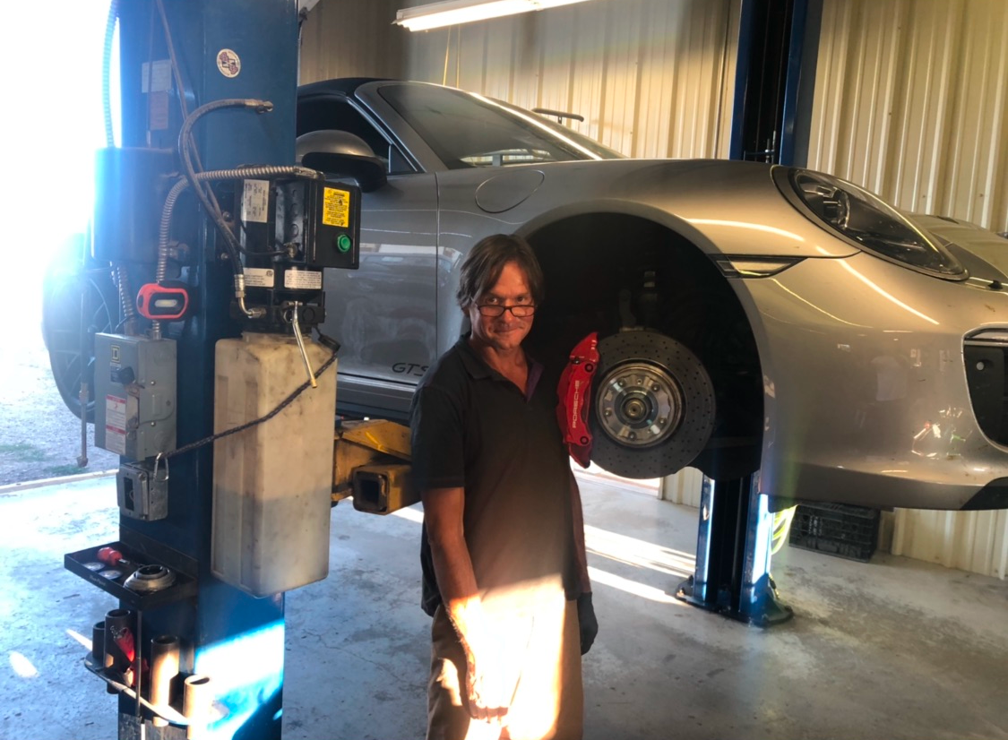 Man smiling next to a silver car lifted on a hoist in a garage, with a view of the car's red brake.