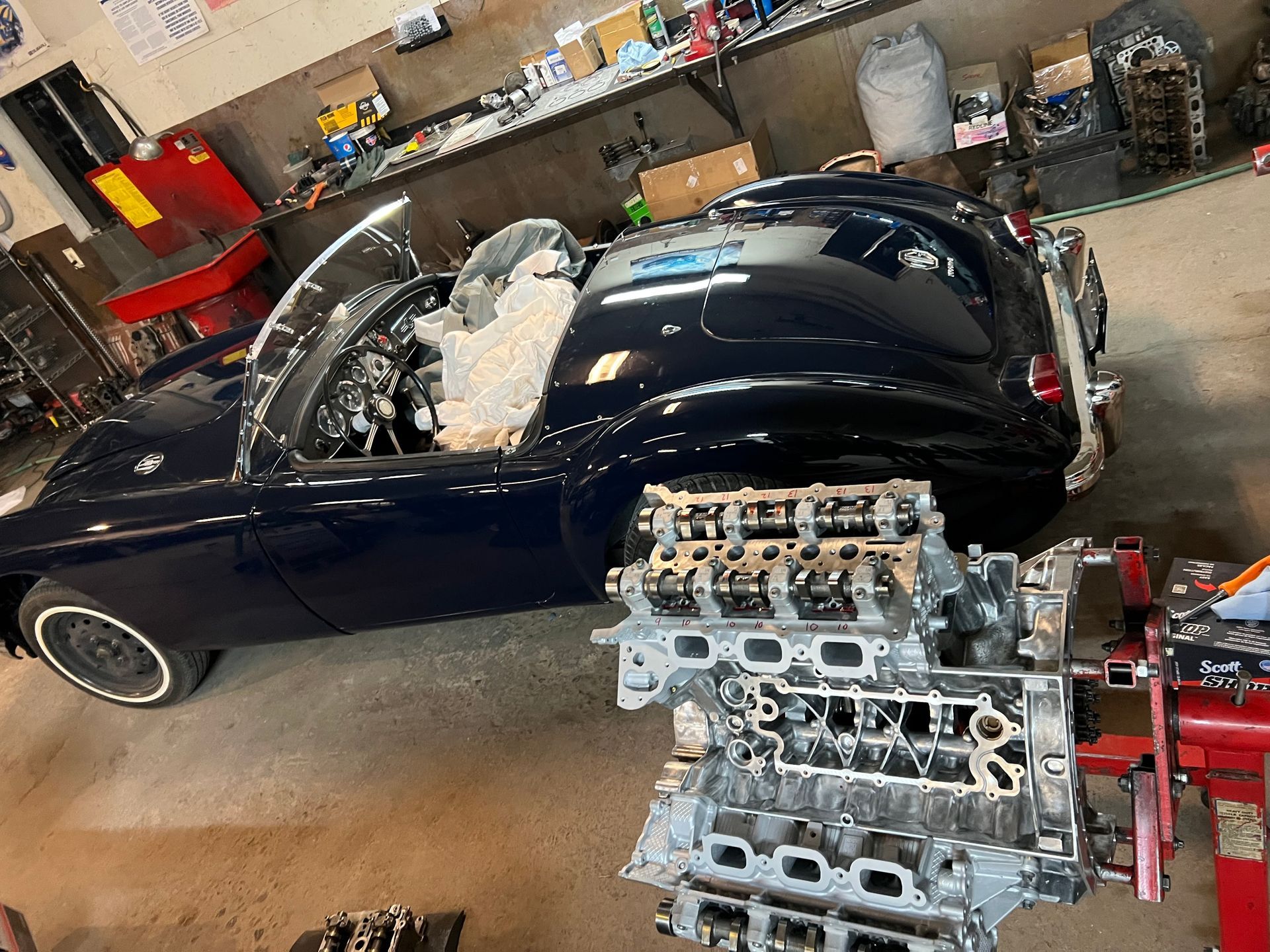 Dark blue convertible car in a garage next to a disassembled engine.
