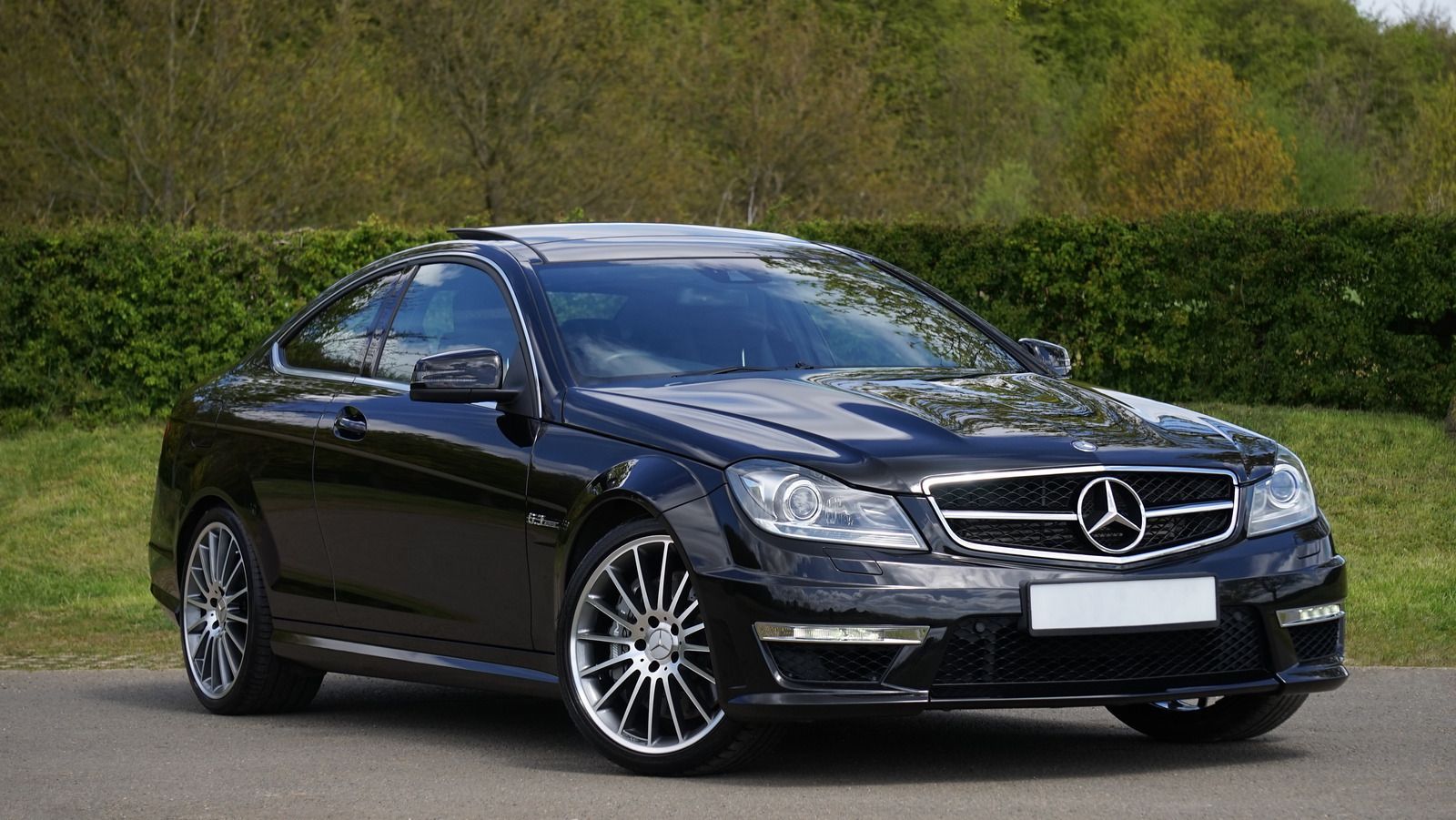 Black Mercedes-Benz coupe with silver wheels parked on a paved surface with green grass and trees in the background.