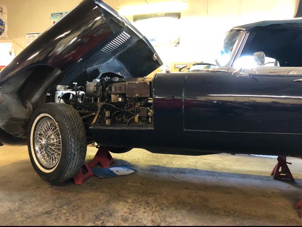 Dark blue Jaguar E-Type with hood open, up on jacks, in a garage.