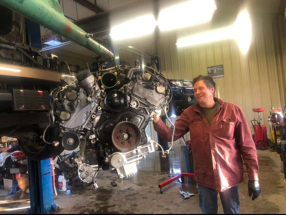 Mechanic with a large engine in a repair shop. Man points at engine, smiling.