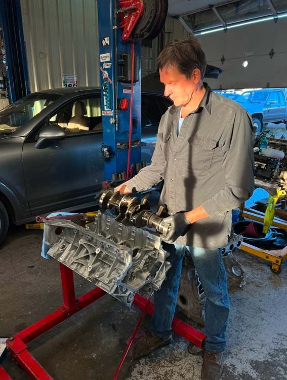 Mechanic inspecting engine block in a garage. He's wearing a grey shirt and jeans. A car is visible in the background.
