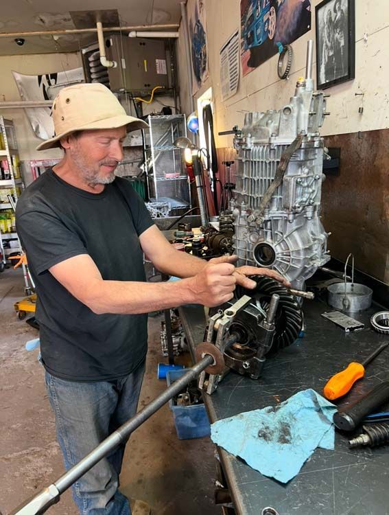 Mechanic points to gear, working on car part. Wearing hat, in garage setting.