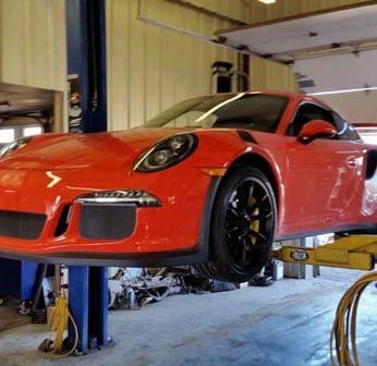 Bright orange Porsche 911 GT3 RS on a lift in a repair shop, black wheels, yellow lift arms.