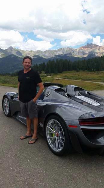 Man in casual wear standing by a gray Porsche 918 Spyder roadster, mountains in the background.