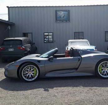 A gray Porsche 918 Spyder convertible parked in front of a building with a black SUV.