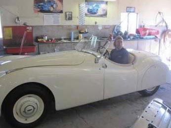 Man sitting in a white vintage Jaguar car inside a garage.