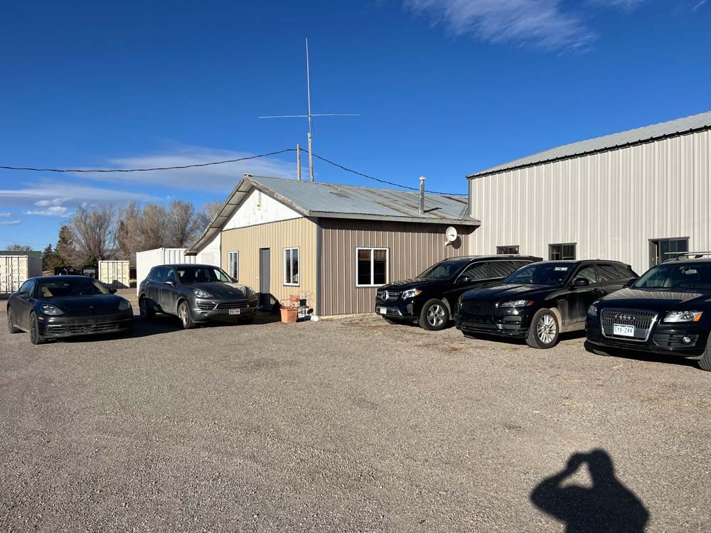 Cars parked in front of a building with antenna on roof, sunny day, gravel lot.