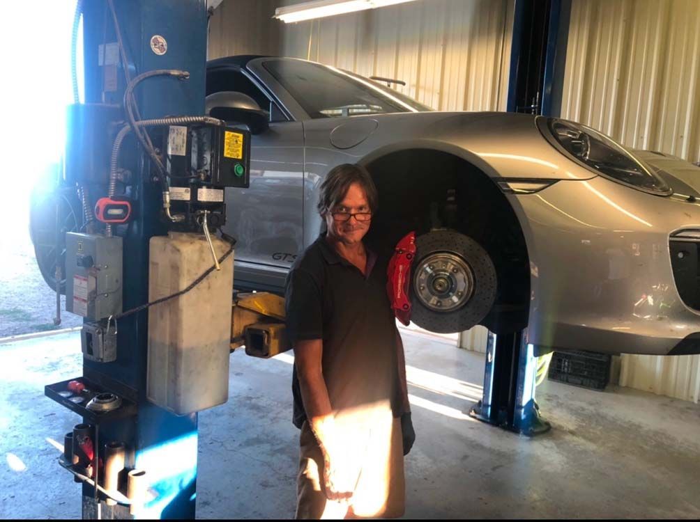 Mechanic beside a silver Porsche lifted in a garage. The car's wheel is removed, showing red brake calipers.