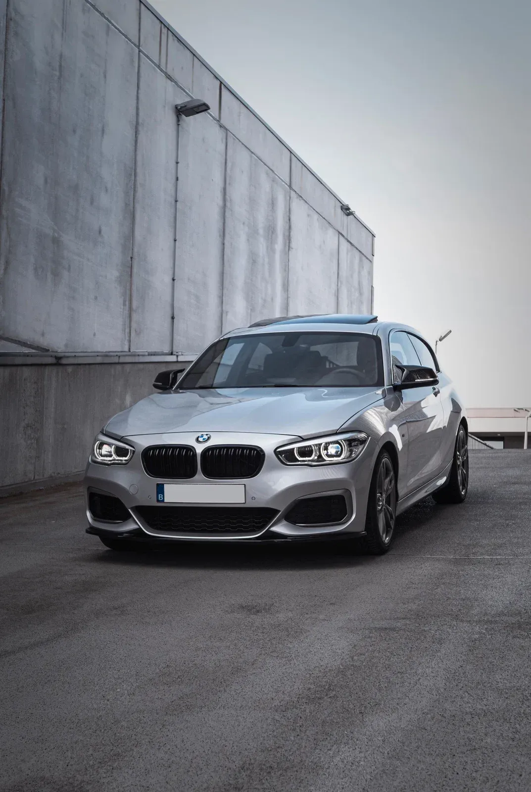 Silver BMW hatchback parked in front of a concrete wall, on an asphalt surface.