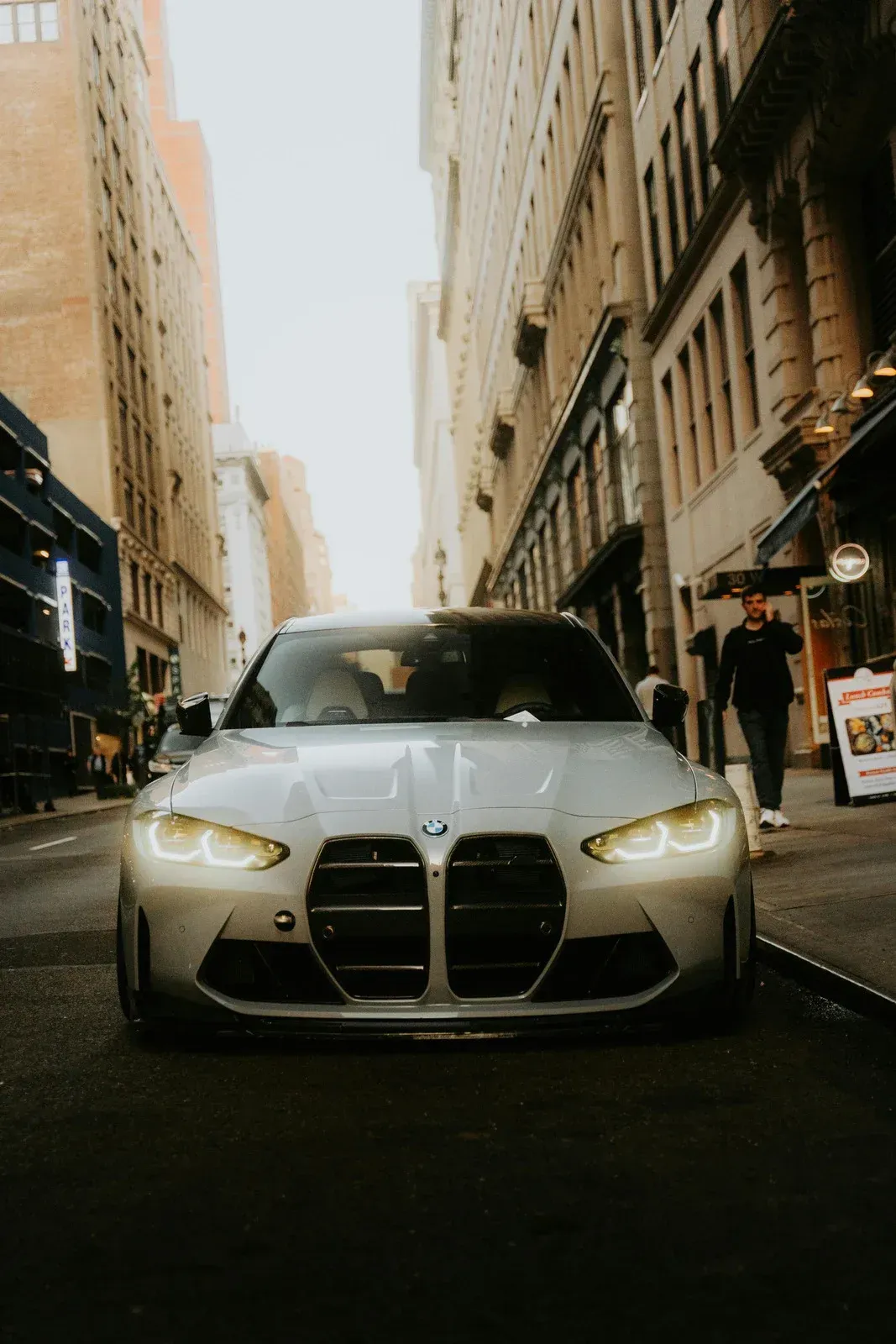 Silver BMW car in narrow city street, headlights on, buildings on either side.