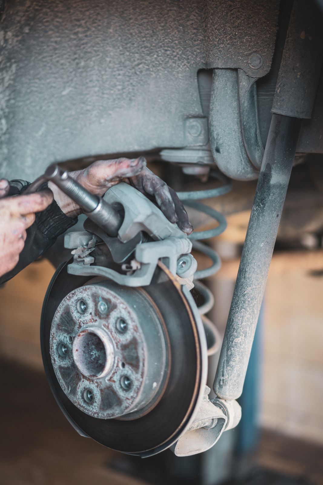 Hands working on a car's brake system; rotor, caliper, and suspension components are visible.