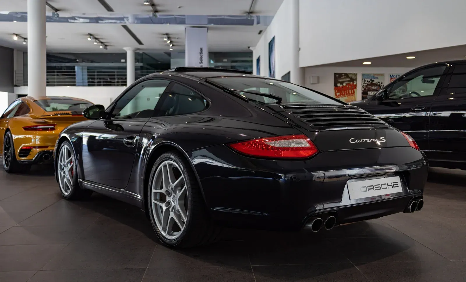 Black Porsche 911 Carrera in a showroom. Another car in yellow to the left.