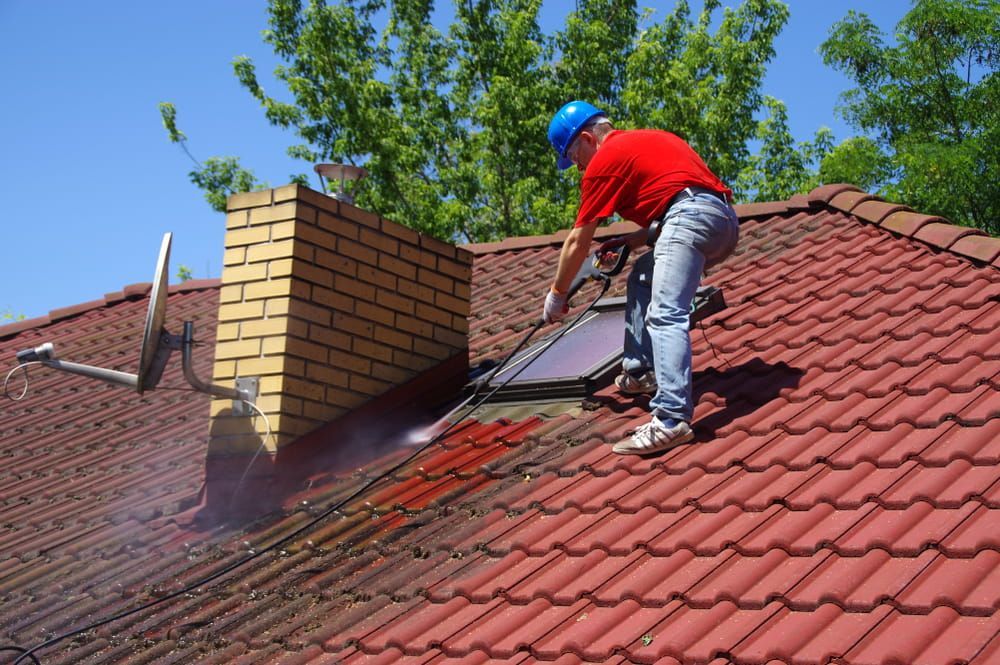 A Man in a Red Shirt is Cleaning the Roof of a House — Roof Restoration Masters In Central Coast, NSW
