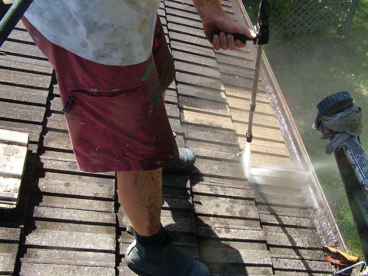 A Man is Cleaning the Roof of a House With a High Pressure Washer — Roof Restoration Masters In Morisset Park, NSW