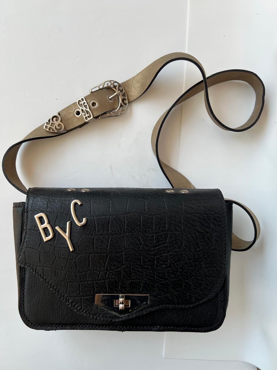 Bolso bandolera de piel negra con hebilla plateada y letras "BYC".