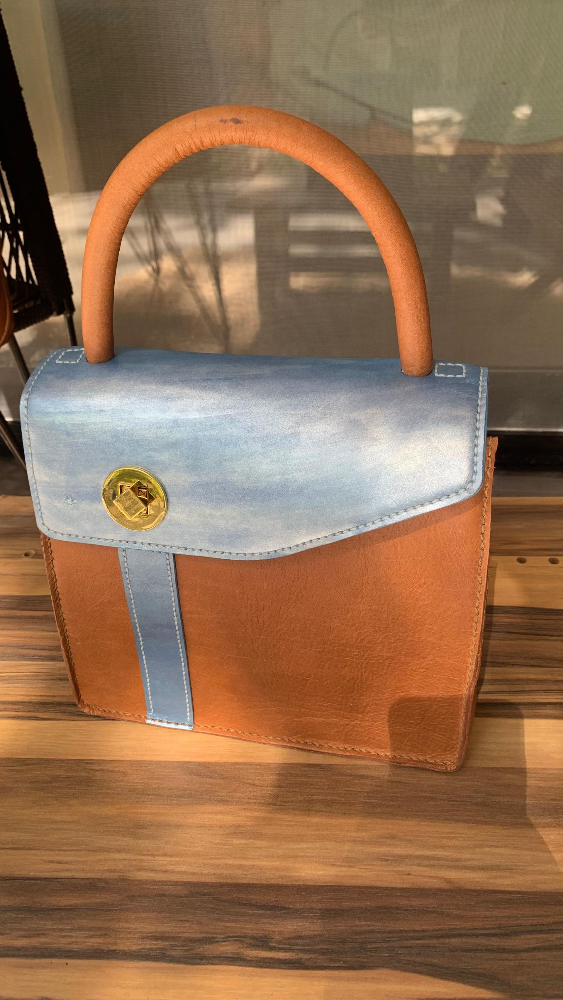 Bolso de mano de color marrón y azul con asa redondeada de color marrón, apoyado sobre una superficie de madera.