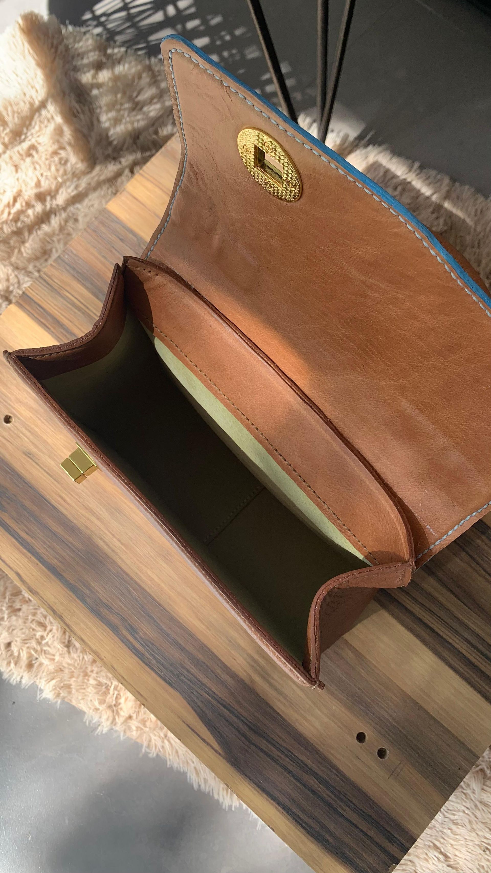 Bolso de mano de color marrón y azul con asa redondeada de color marrón, apoyado sobre una superficie de madera.