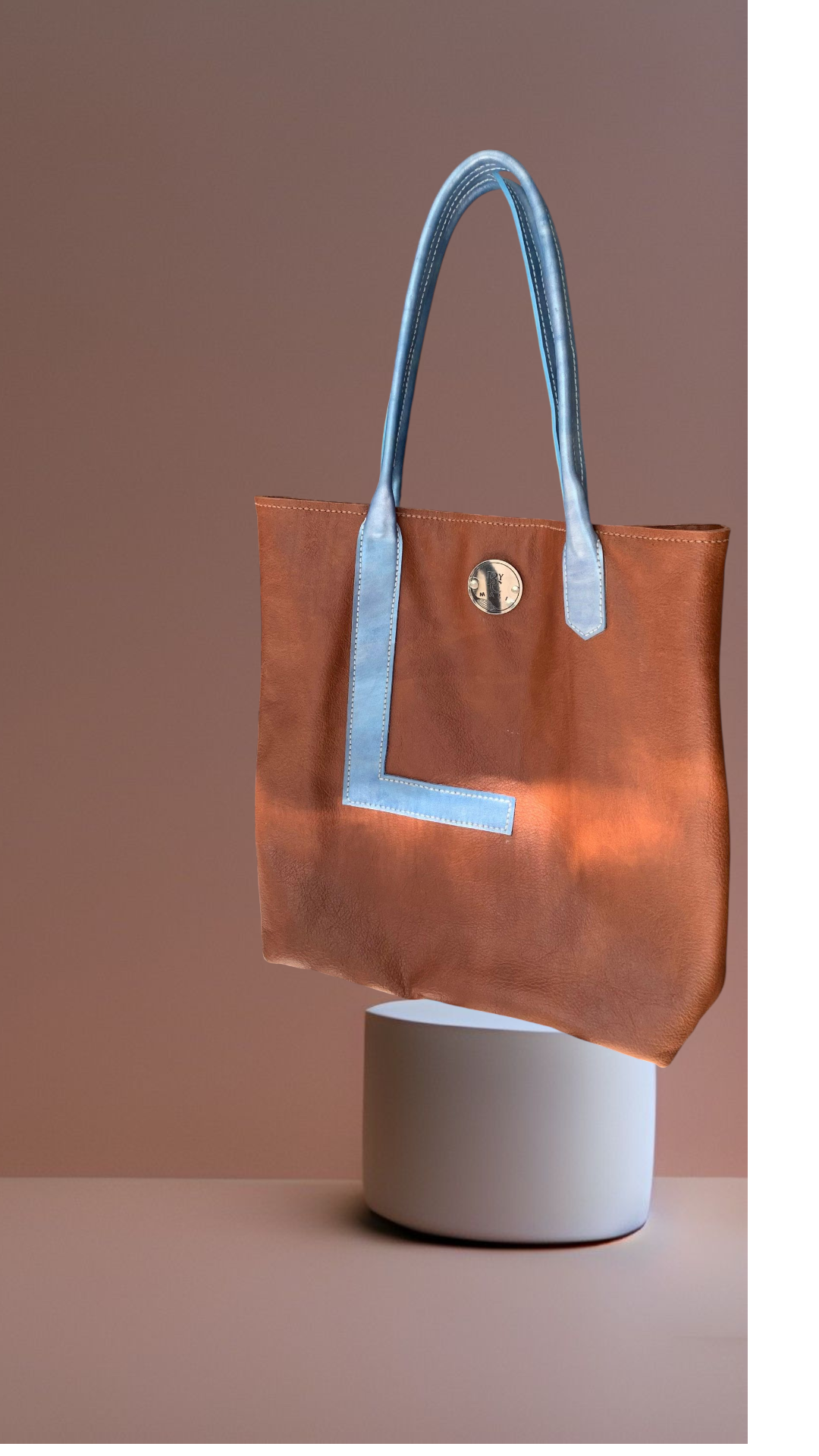 Bolso tote de piel marrón con asas azules, sobre un pedestal blanco y sobre un fondo rosa.