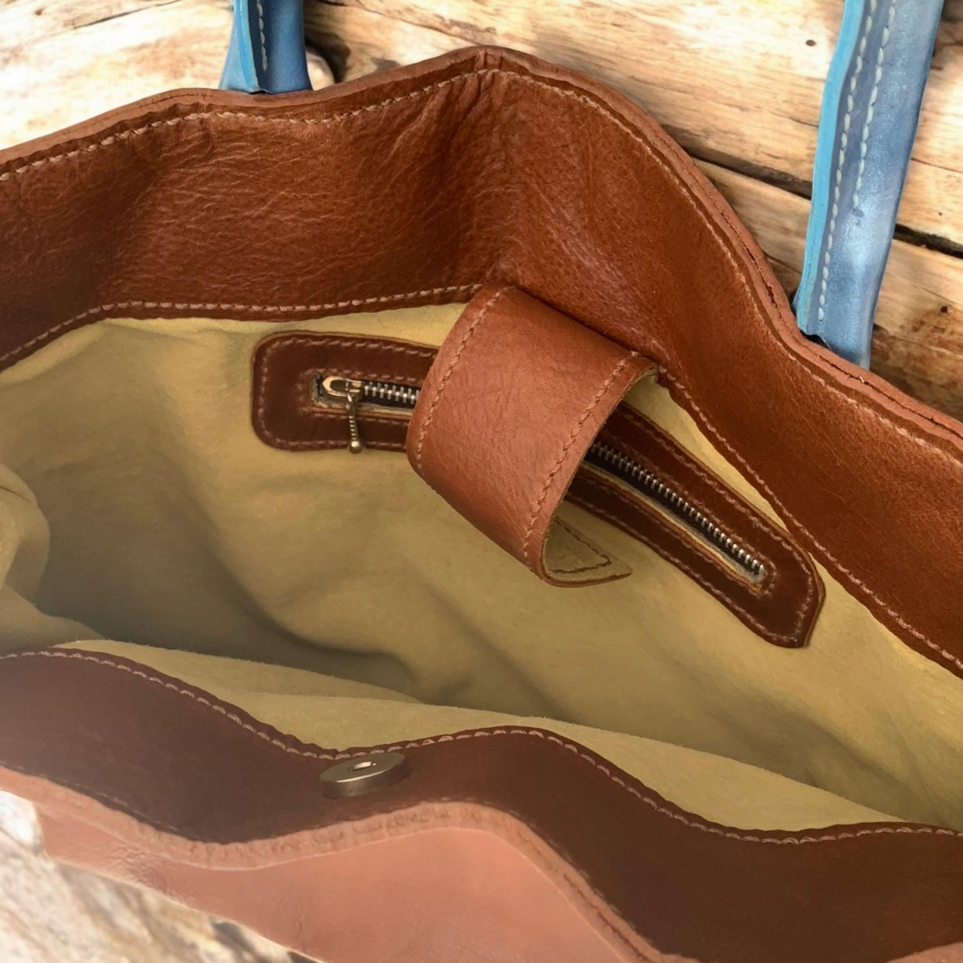 Bolso tote de piel marrón con asas azules, sobre un pedestal blanco y sobre un fondo rosa.