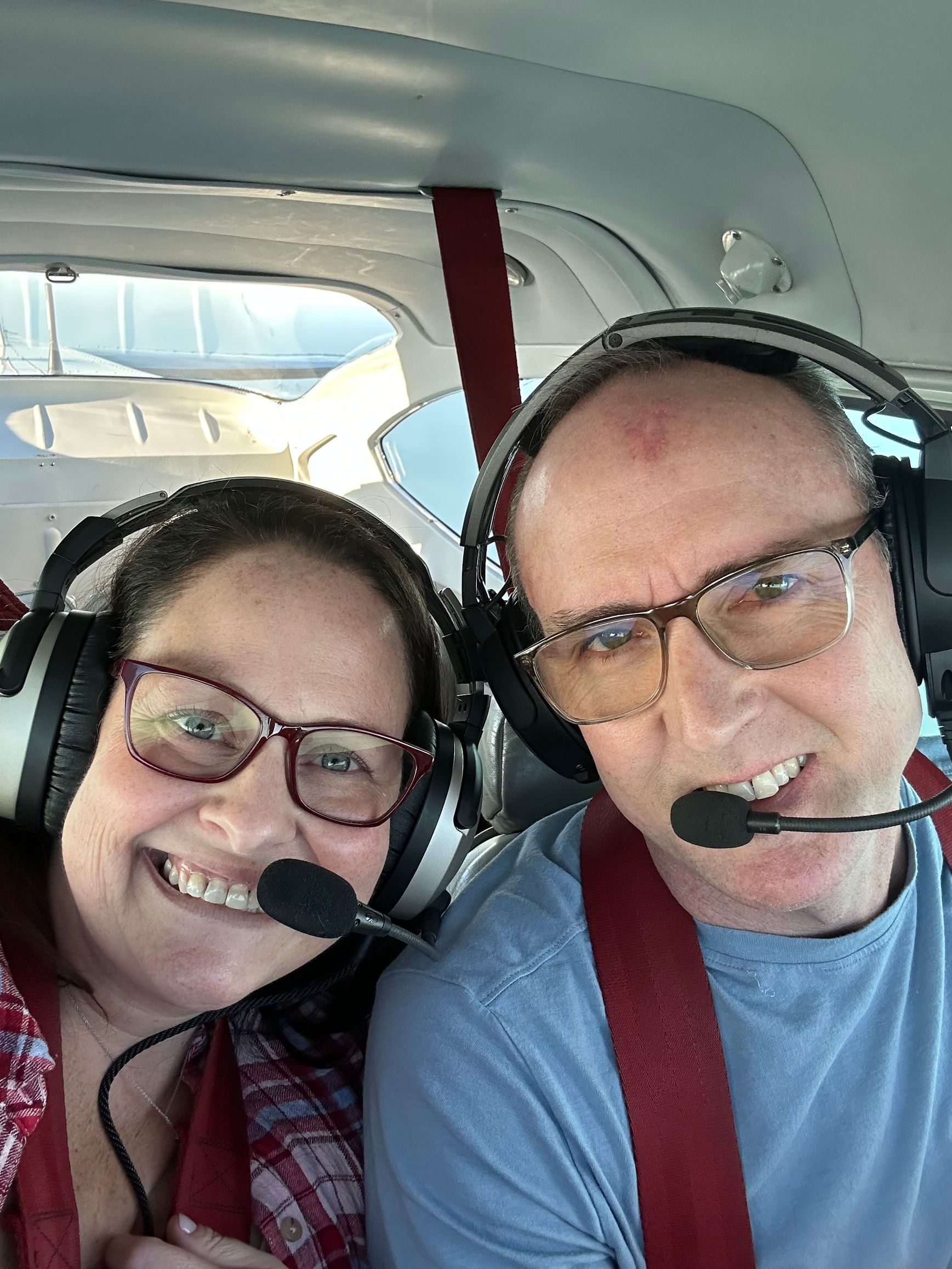 Two people in a small plane cockpit wearing headsets and glasses, smiling. Red seatbelts visible.