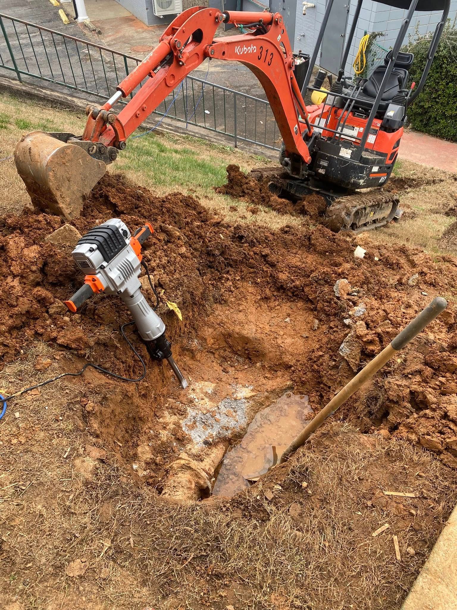 Orange mini excavator digging a trench beside a silver jackhammer in a mulch-covered yard