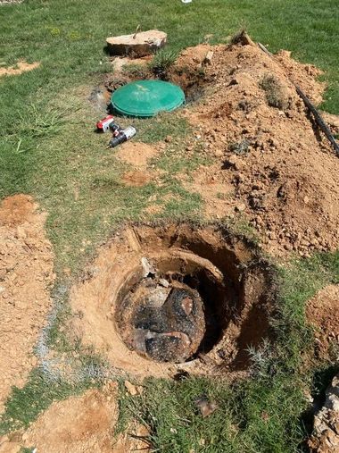 Open dirt hole in a grassy yard beside a mound of soil and a green lid