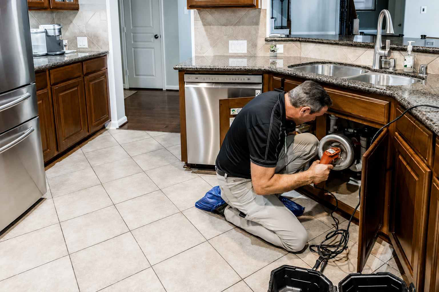 Technician repairing a dishwasher in a kitchen, kneeling beside the open appliance with tools nearby.