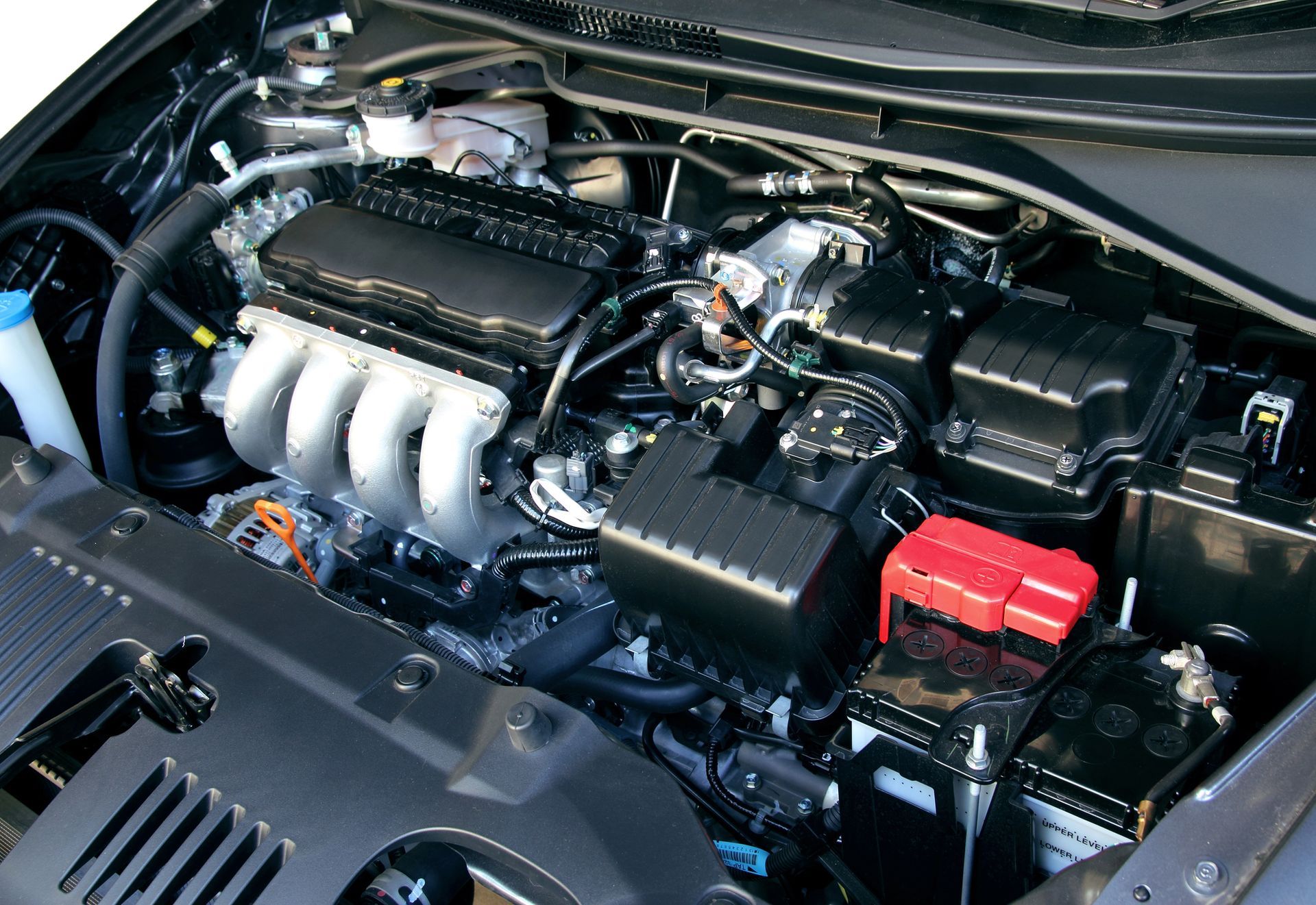 Vano motore dell'auto con componenti neri, collettore di aspirazione argentato e terminale della batteria rosso in vista.