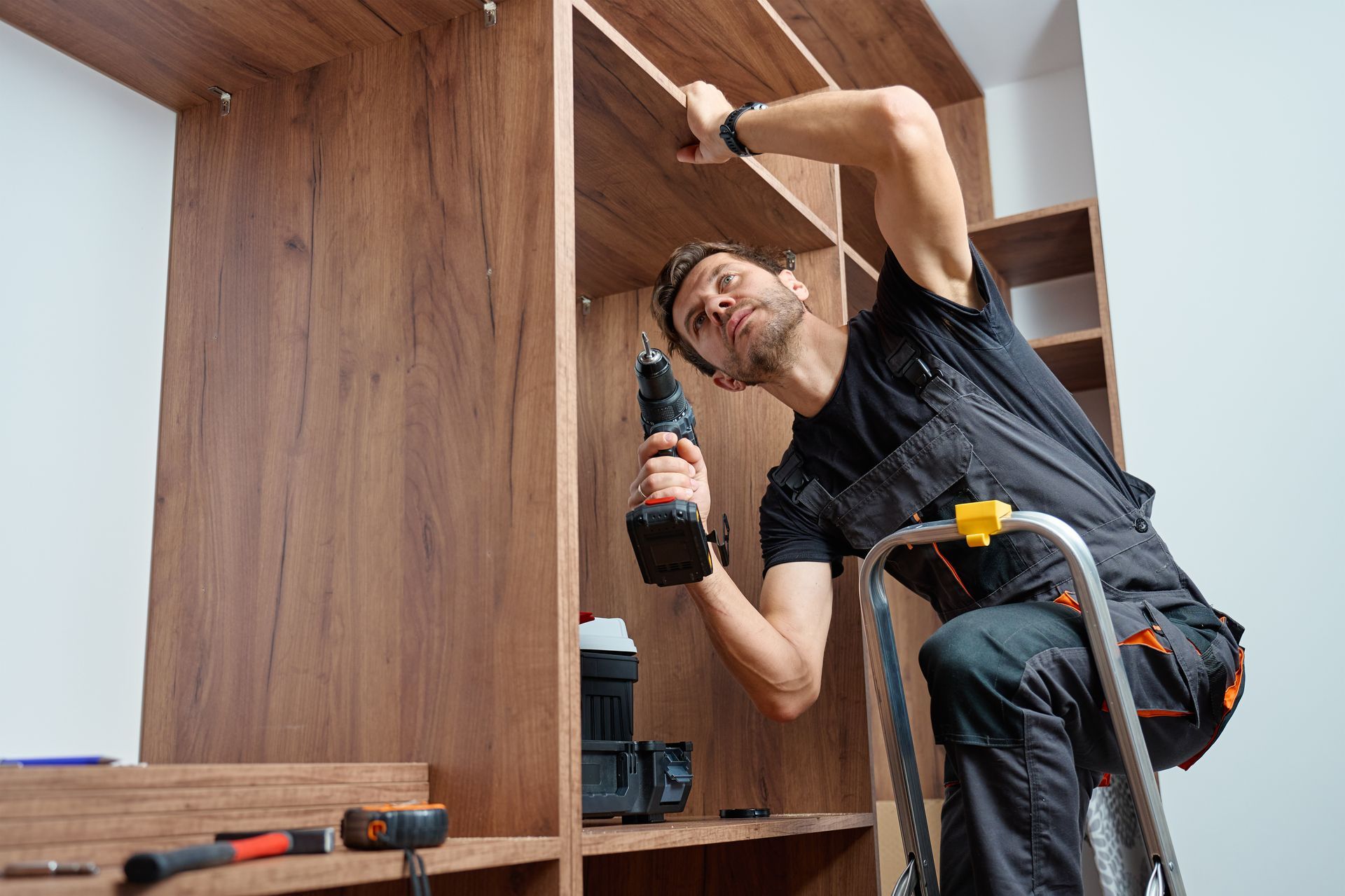 Man on ladder installing wooden shelving with a drill.