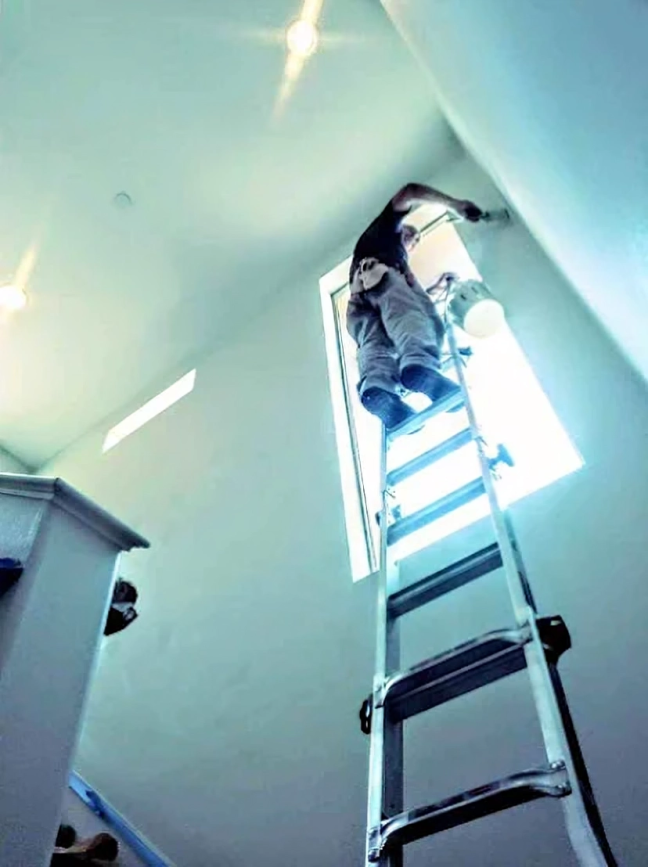 Person on ladder painting a high wall near a window. Bright, indoor setting.