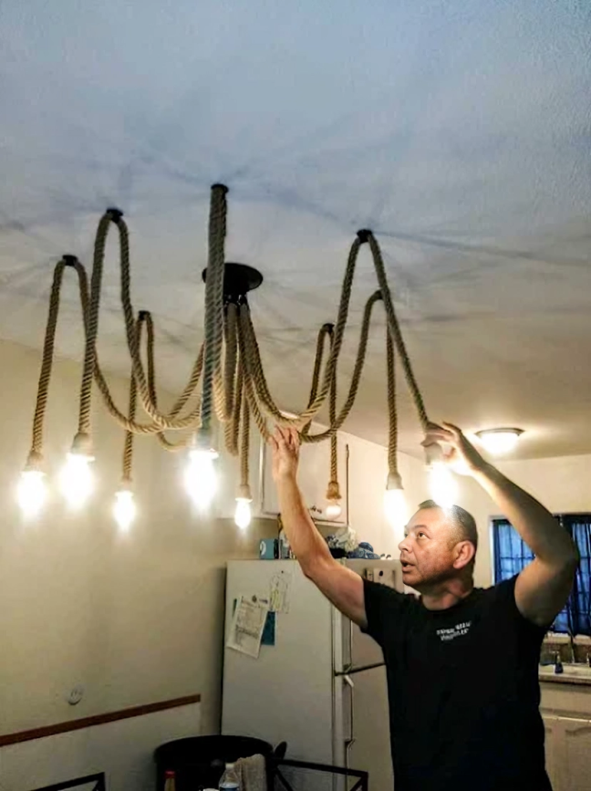 Man installing a rope chandelier in a kitchen. The light fixture is made of rope with lit bulbs.