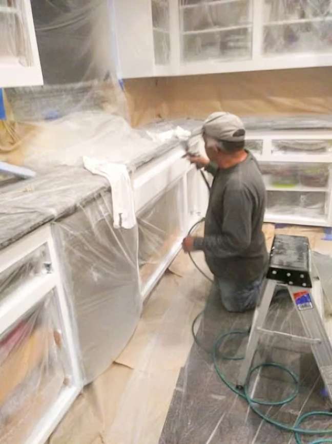 Man spraying white paint on kitchen cabinets; he is kneeling.