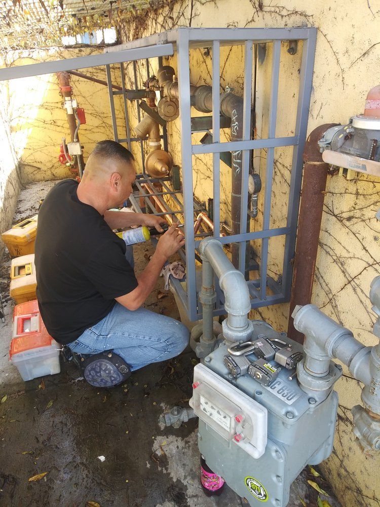 Man in black shirt working on pipes near a gas meter; outdoors, utility area.
