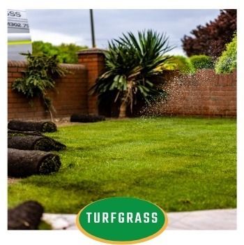 Stacks of artificial turf on a green lawn in a backyard with brick wall and plants, labeled TURFGRASS