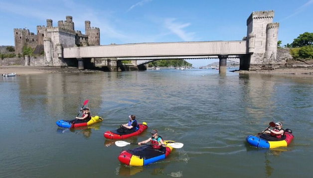 Four brightly coloured packrafts being paddled in front of an ancient castle with a bridge across an estuary