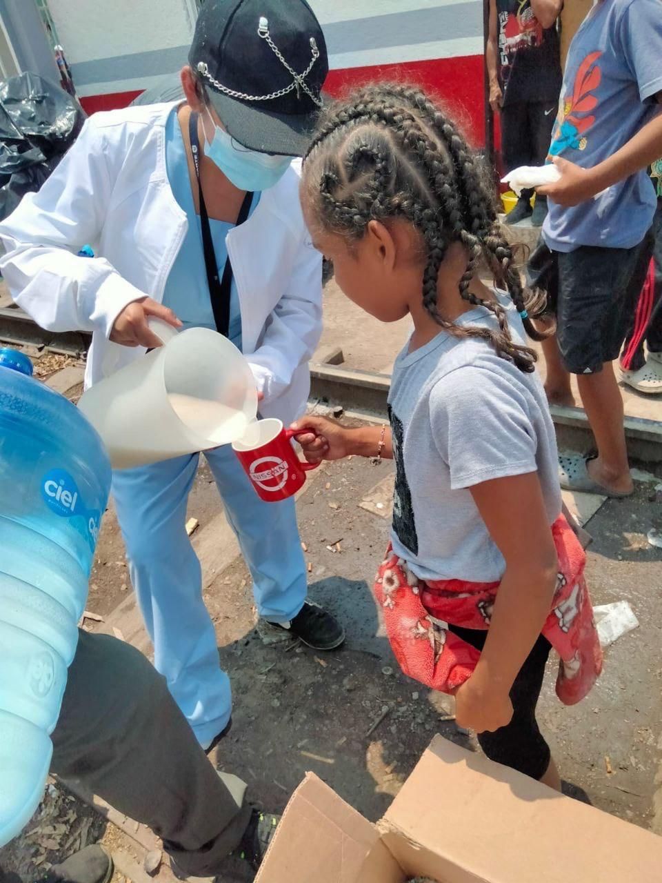 Una persona con uniforme médico vierte leche en una taza para un niño al aire libre, cerca de un tren.
