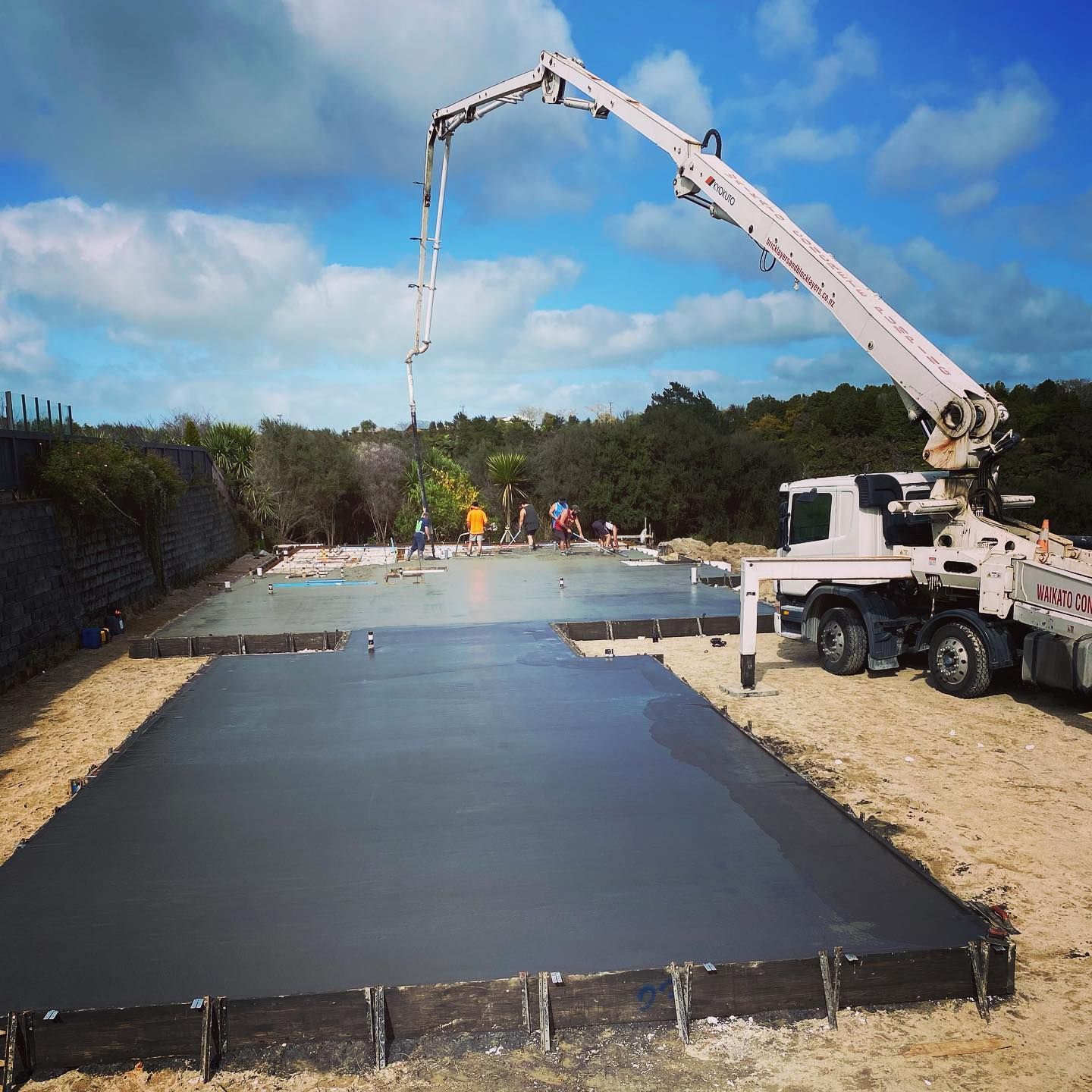 Concrete being poured onto a foundation by a truck-mounted boom; construction workers in the background.